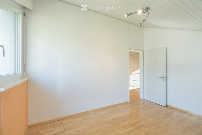 Empty room with light-colored walls, wooden flooring, and a large window with blinds. A door leads to another room with wooden flooring and a visible balcony area.