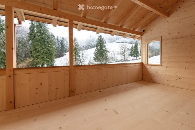 Empty wooden room with exposed beams, large windows overlooking a snowy landscape, and a small arched window on the right side.
