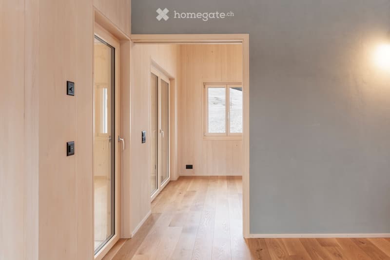 A modern, minimalist hallway with light wooden flooring, light grey walls, and several doors and windows. The space is clean and uncluttered, with natural light coming through the windows.