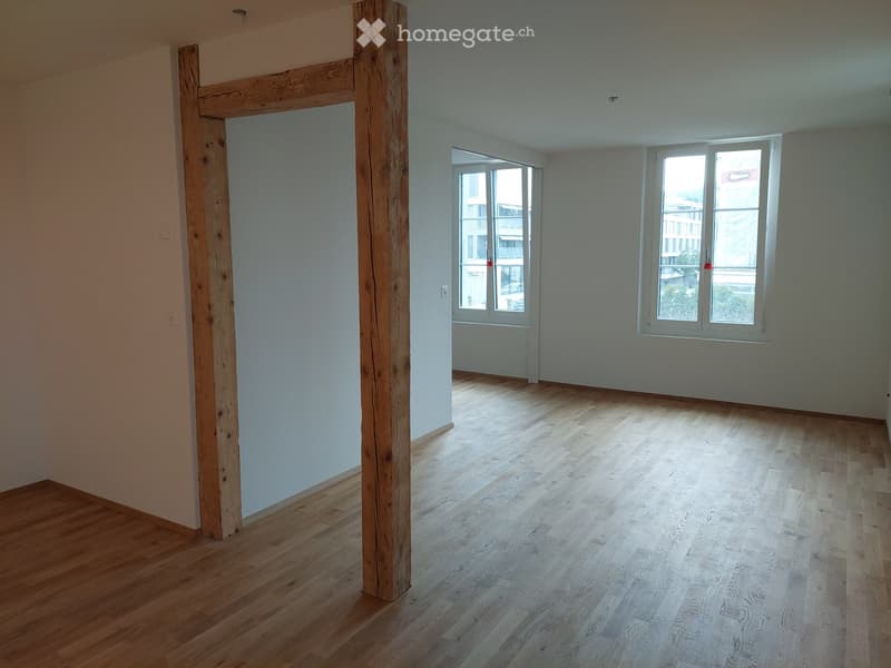 Empty room with wooden flooring, light-colored walls, and large windows with white frames. A wooden structural beam is visible on the left side.