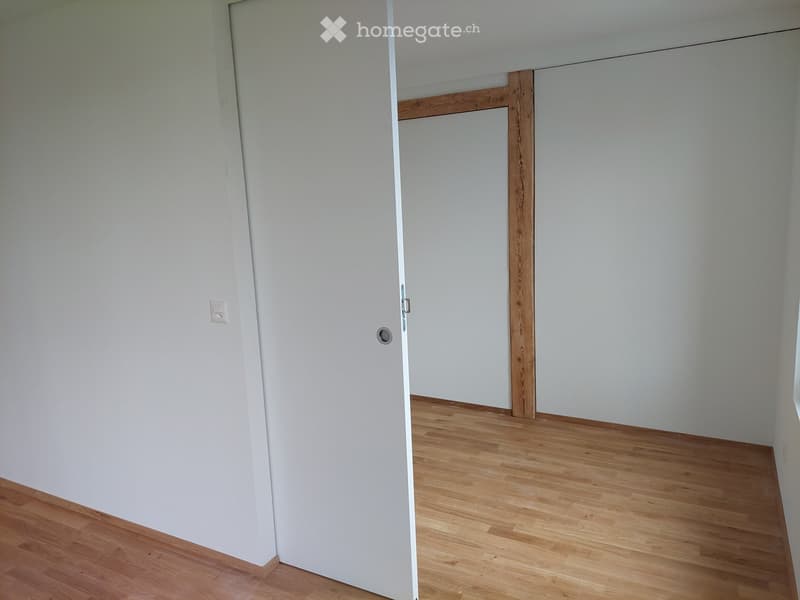 Empty room with light wooden flooring and white walls, featuring a closed white door and a visible electrical outlet.