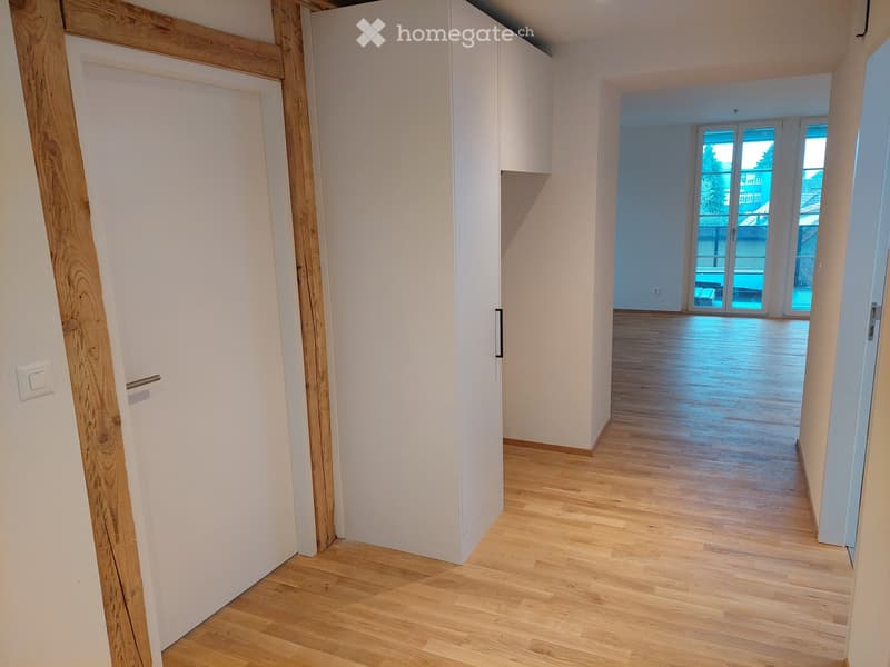 Empty hallway with wooden flooring, white walls, and a door leading to a room with large windows and a balcony view.