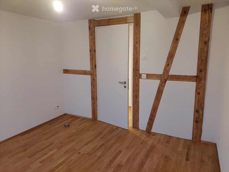 Empty room with wooden flooring, white walls, and a white door leading to another space. Exposed wooden beams frame the doorway.