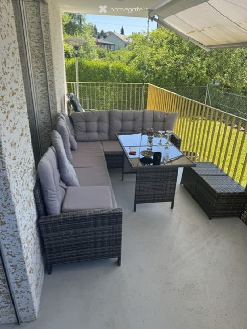 Balcony with a grey sectional sofa, glass coffee table, and wicker storage bench. The balcony has a metal railing and overlooks a green yard.