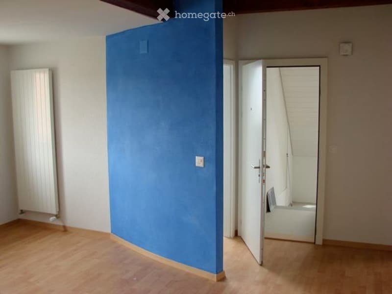 Empty room with wooden flooring, white walls, a blue partition, and an open doorway leading to a small bathroom.