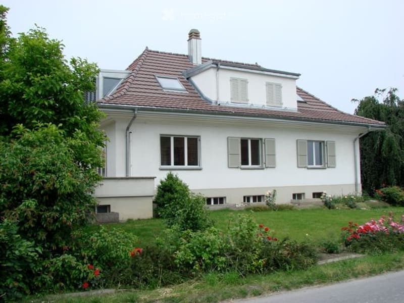 Two-story white house with red-tiled roof, multiple windows with shutters, surrounded by a well-maintained garden with various plants and flowers.