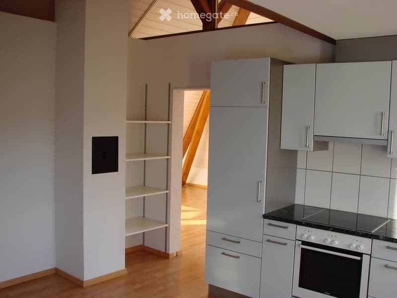 Modern kitchen with light grey cabinets, a stove, an oven, and a shelf unit. The space features wooden flooring and a slanted ceiling with wooden beams.