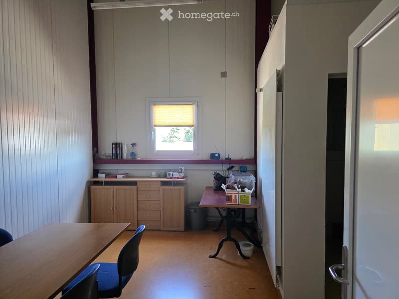 A small kitchen area with a wooden table, chairs, cabinets, and a refrigerator. There is a window with blinds and a small table with various items on it.