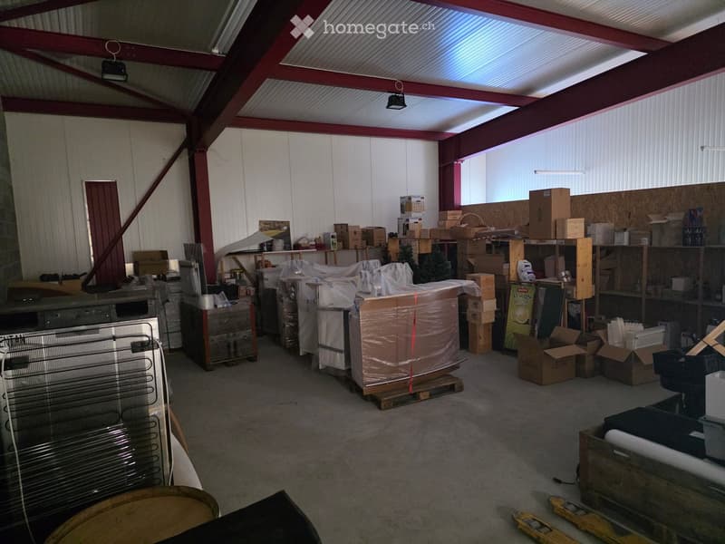 An empty warehouse or storage space with exposed metal beams, corrugated metal walls, and scattered boxes and items, including a large wrapped object, shelves, and a fan.