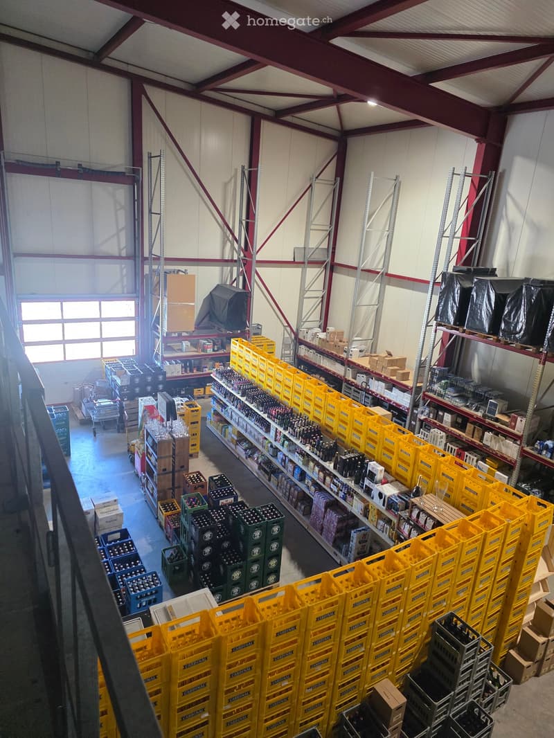 Warehouse with high ceilings, metal shelving units, and numerous yellow crates stacked on the floor. Shelves hold bottles and boxes, with a large window allowing natural light to enter.
