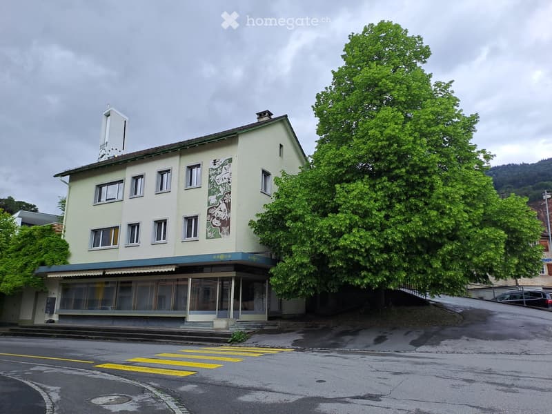 Two-story building with light yellow facade, multiple windows, and a mural on one side. The building has a small awning over the entrance and is surrounded by trees and a road with a crosswalk.