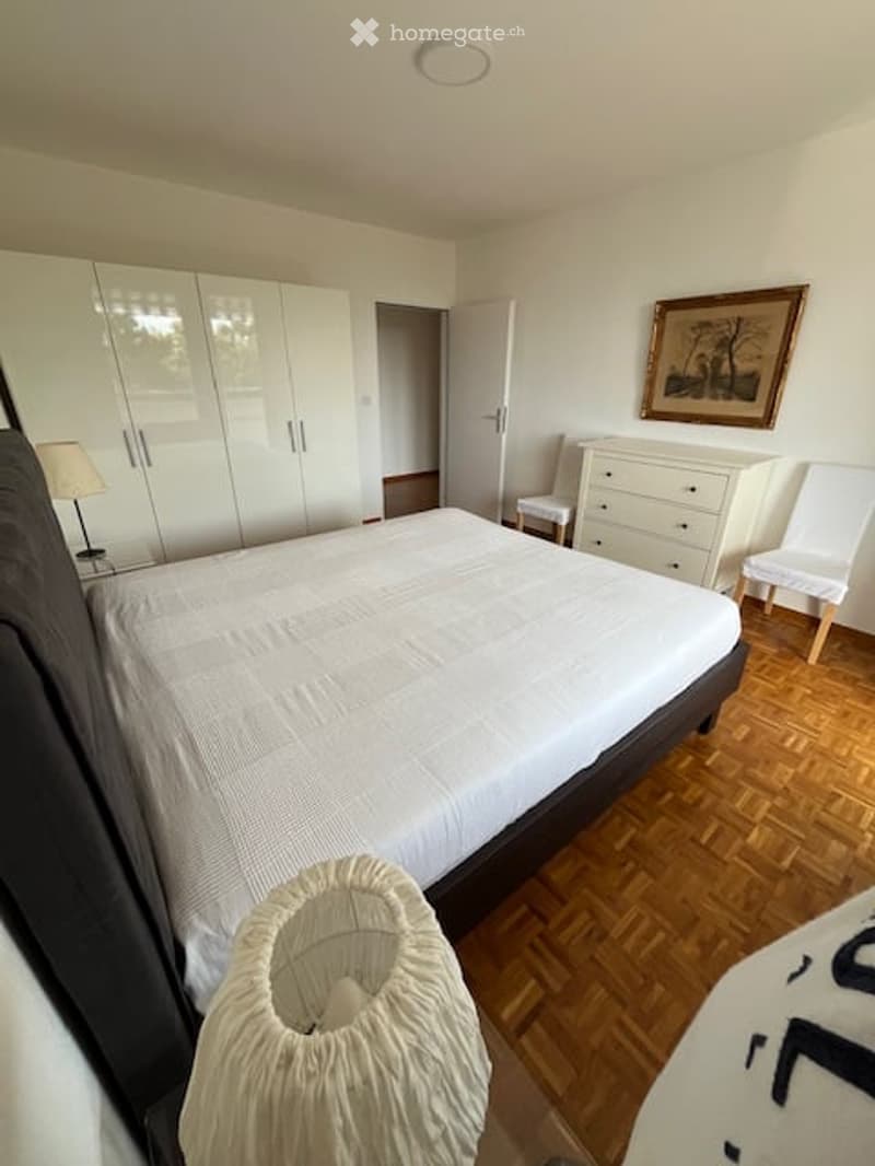 Bright bedroom with wooden flooring, large bed, white wardrobe, two chairs, a table lamp, and a framed picture on the wall.