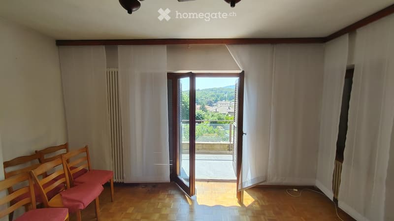 Empty room with wooden flooring, large window with white curtains, and a set of wooden chairs with red cushions near the window.