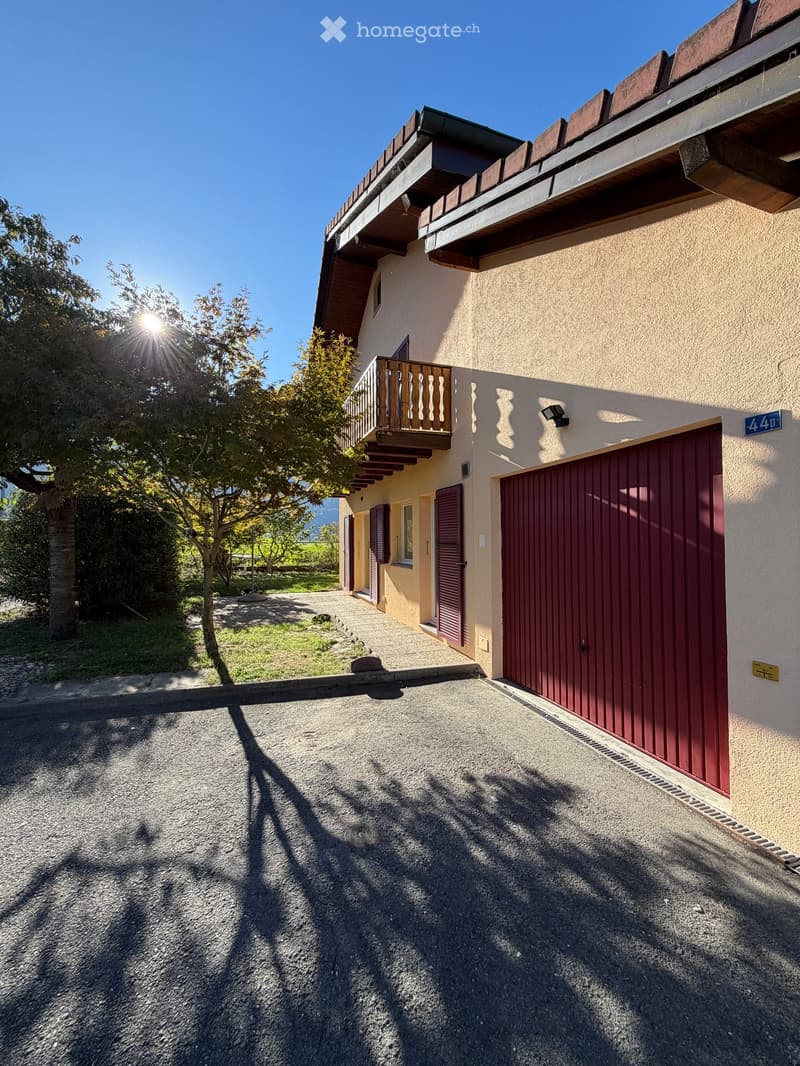 Two-story house with beige walls, red garage door, and balcony with wooden railing. The driveway is paved, and there is a tree and greenery in the foreground.