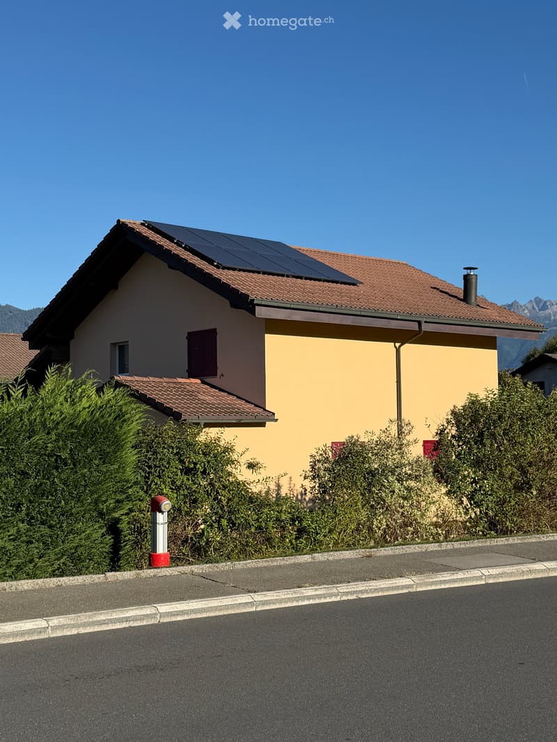 Two-story house with a tiled roof, solar panels, and a chimney. The house has light yellow and beige walls, with a small garden area in front.