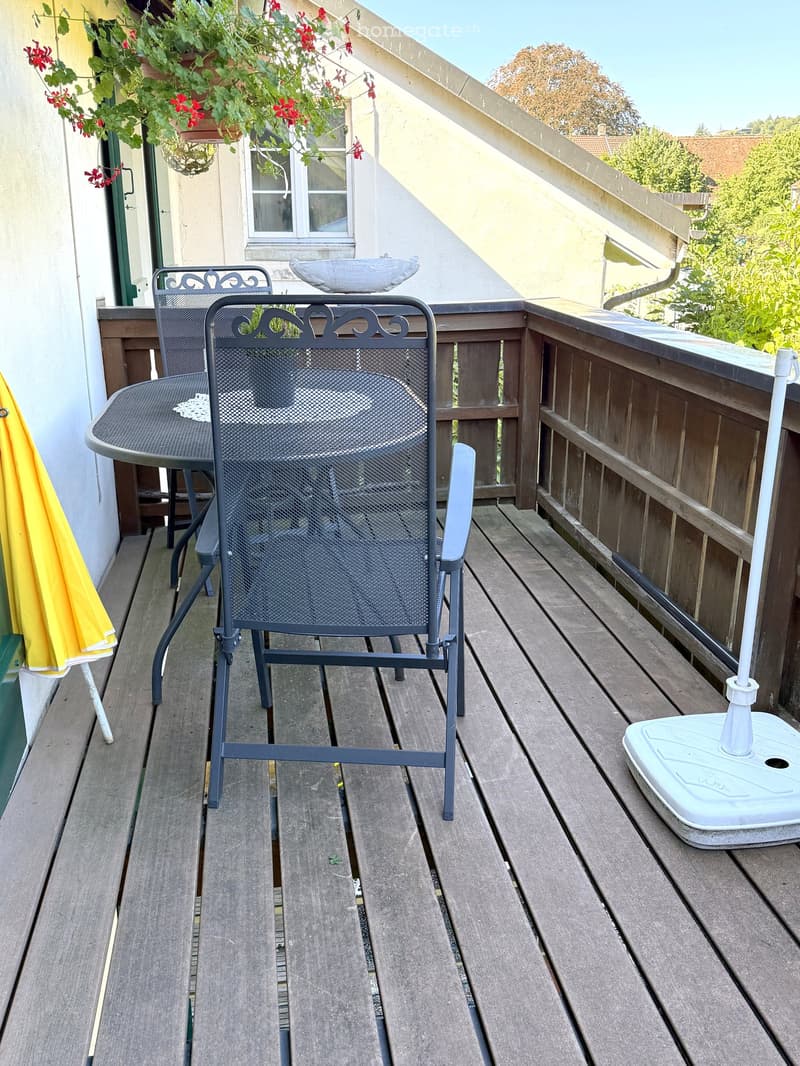 Wooden balcony with a metal chair, small table, and umbrella stand. Potted plants and a decorative bowl are visible in the background.