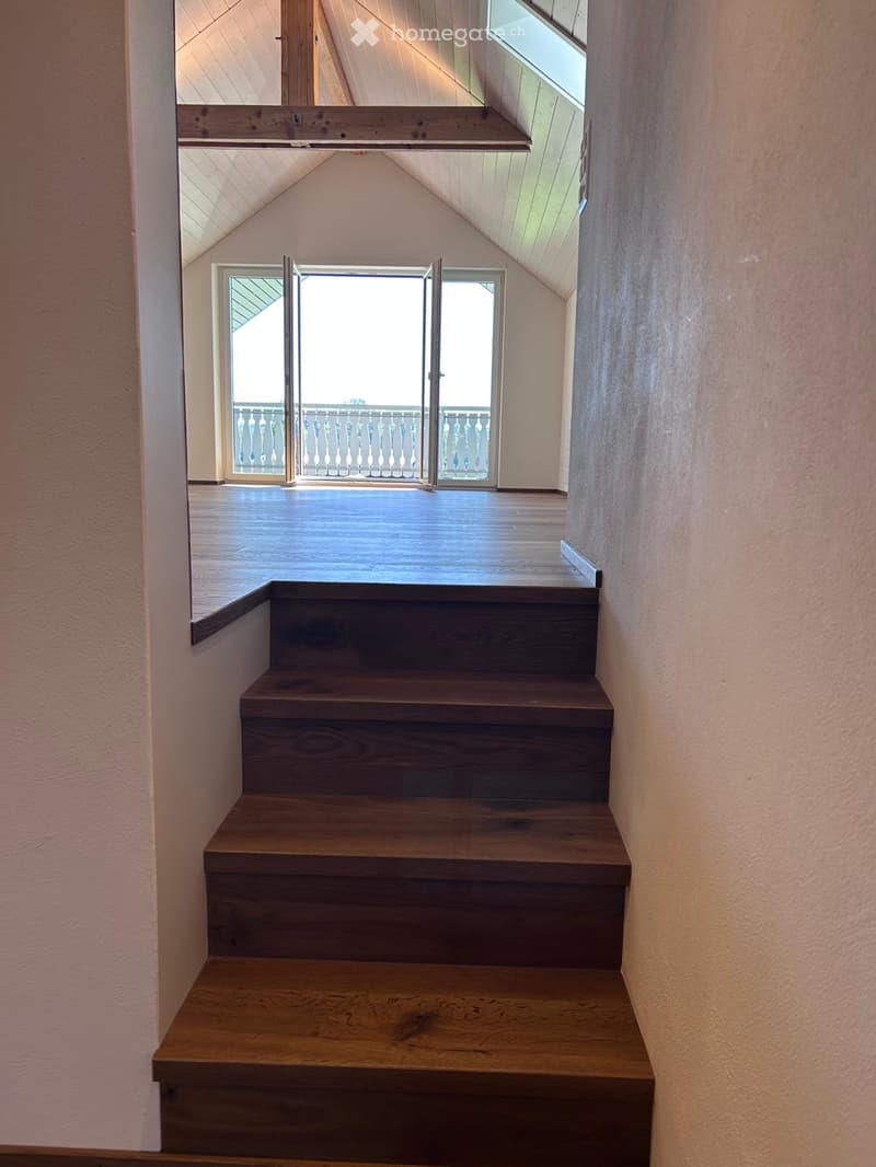 Wooden staircase leading to an open loft area with exposed wooden beams and a skylight window above.