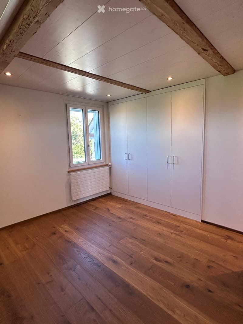 Empty attic room with wooden flooring, white walls, built-in wardrobes, and a window with a radiator below it.