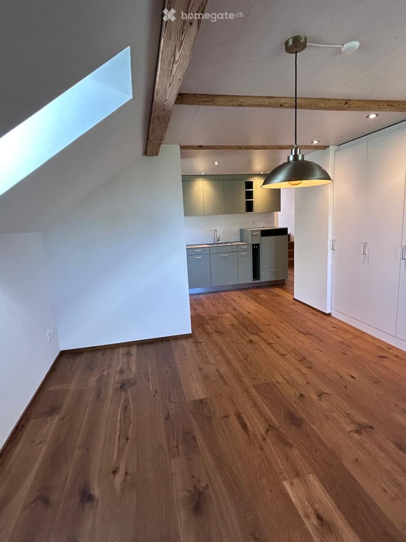 Empty room with wooden flooring, white walls, and a modern kitchen area in the background. No furniture or equipment is present.