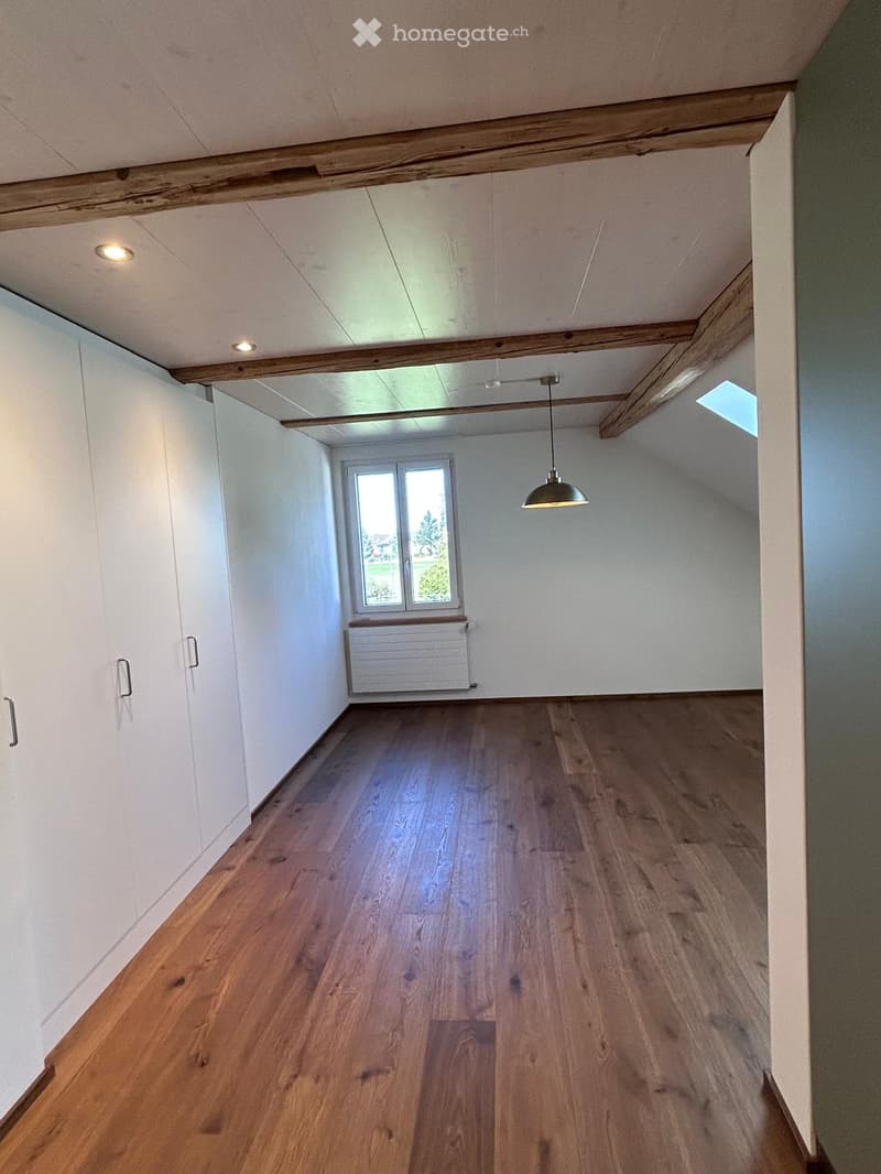 Empty room with wooden flooring, white walls, exposed wooden beams on the ceiling, and a single pendant light fixture. There is a window with a radiator below it and built-in white cabinets along one wall.