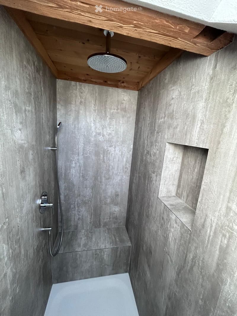 Modern bathroom with grey stone walls, a sleek showerhead mounted on a wooden shelf, and a built-in niche for storage.