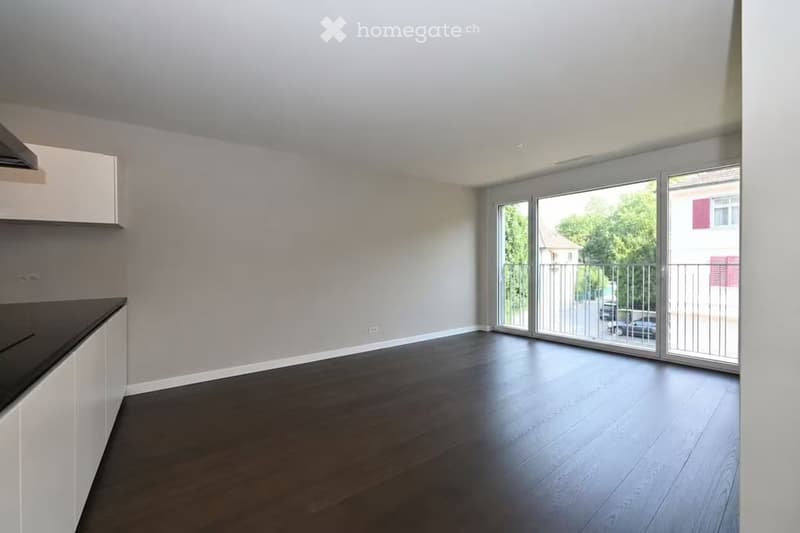 Empty room with light grey walls, dark wooden flooring, and a white kitchen cabinet visible on the left side. Large sliding glass doors lead to a balcony with a metal railing.