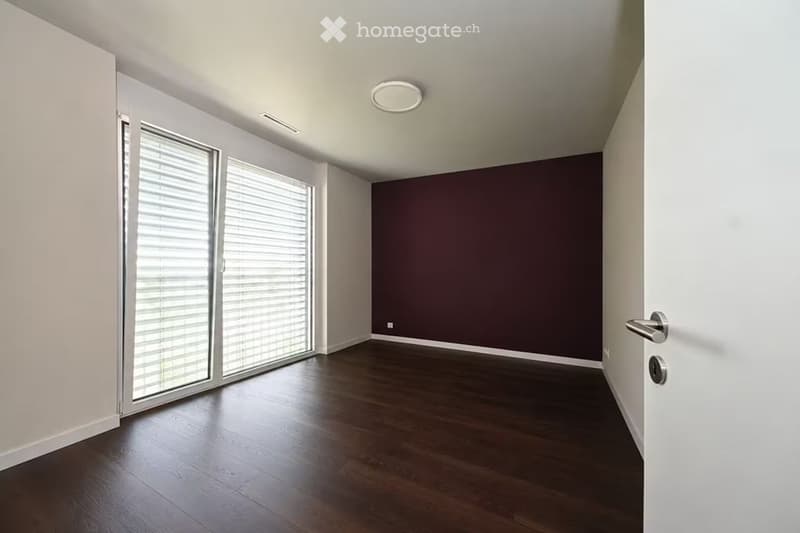 Empty room with dark wooden flooring, white walls, large window with blinds, and a door.
