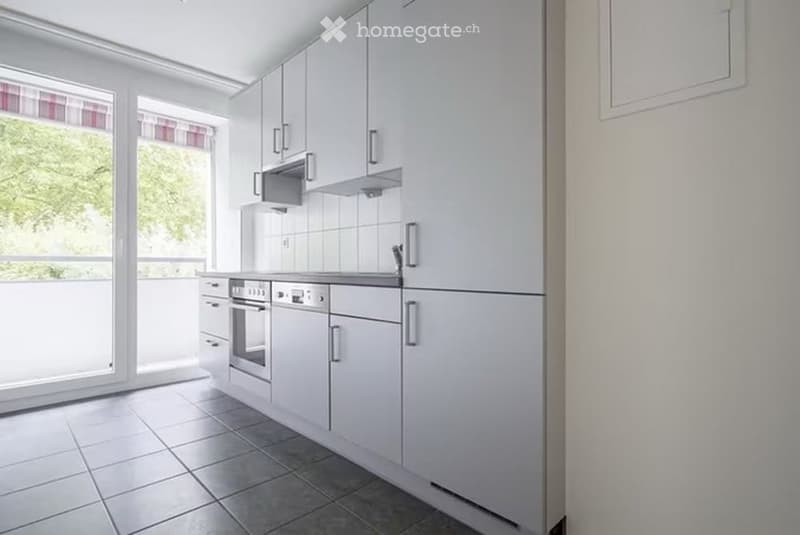 Modern kitchen with white cabinets, stainless steel appliances, tiled floor, and large windows offering a view of greenery.