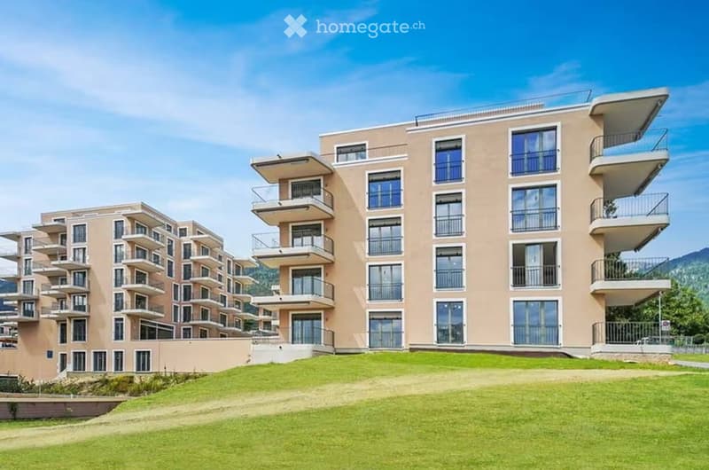 Modern multi-story residential building with balconies and large windows, surrounded by green lawn.