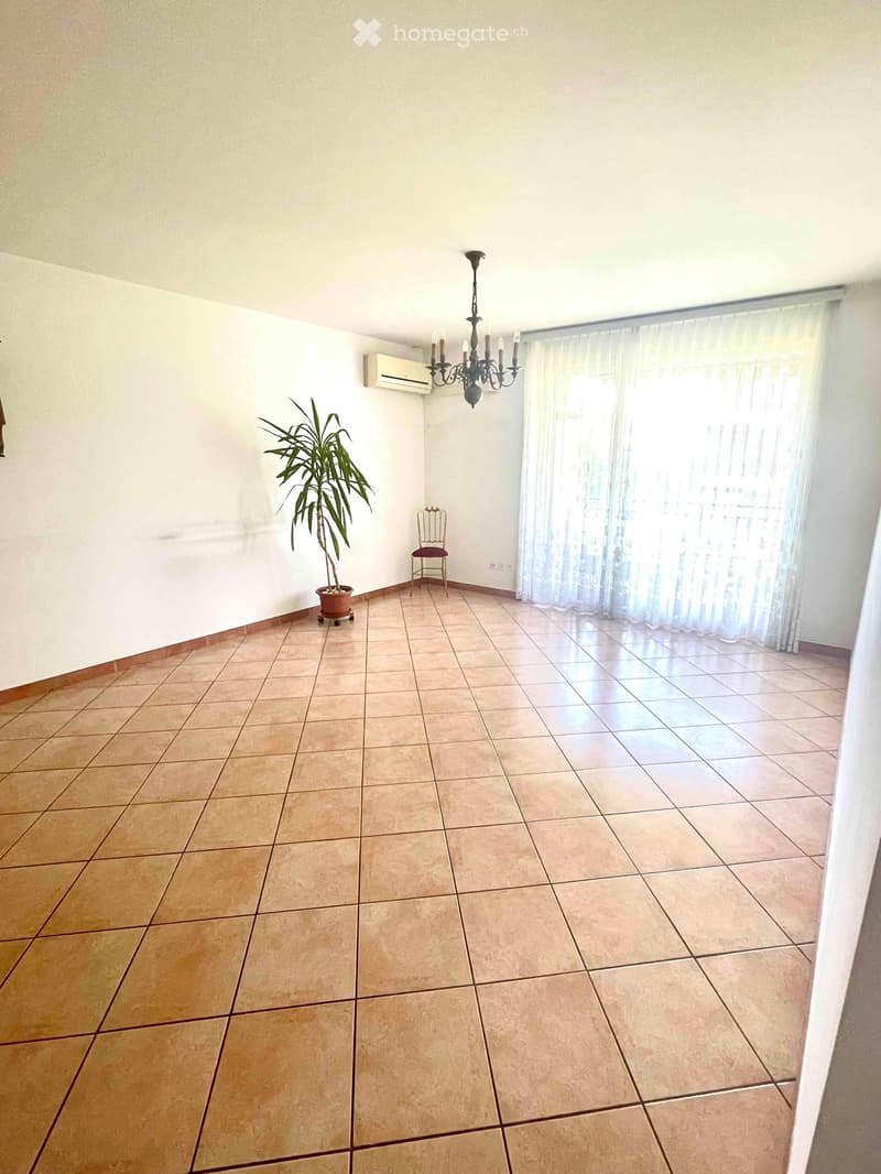 Empty room with tiled floor, white walls, large window with sheer curtains, air conditioning unit, chandelier, small decorative chair, and potted plant.