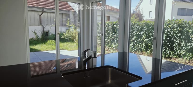 Modern kitchen with large windows overlooking a garden, featuring a stainless steel sink and faucet.