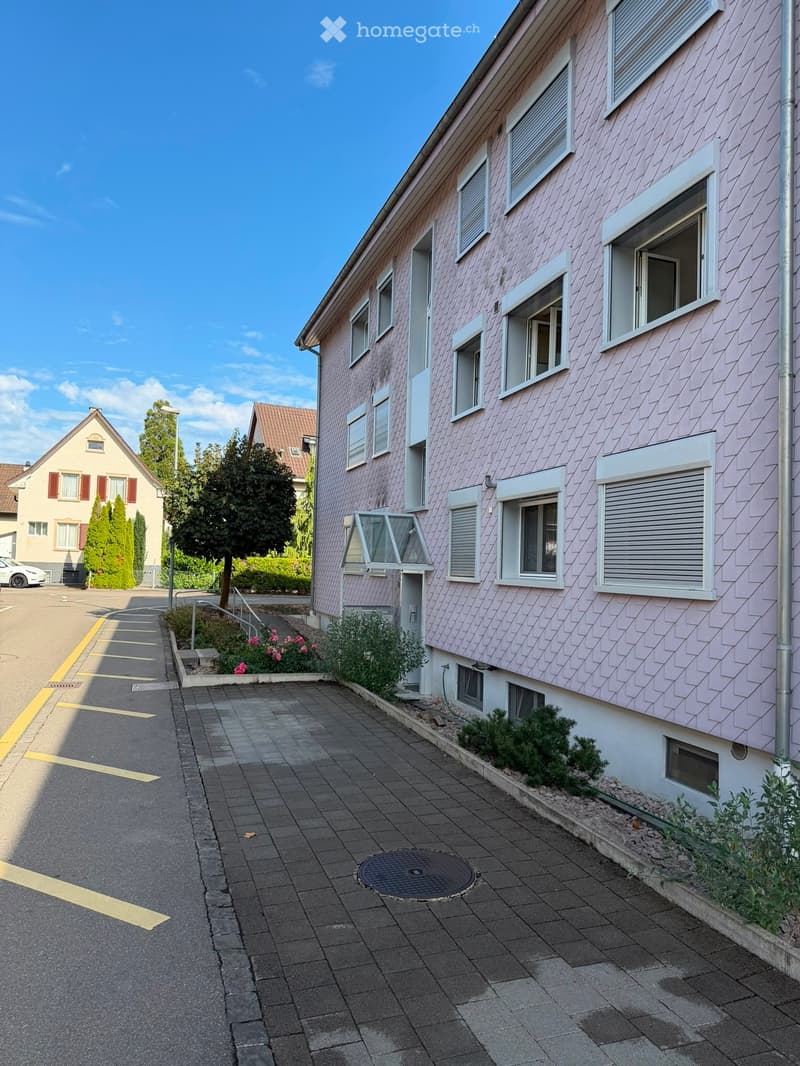 A two-story residential building with pink siding, multiple windows, and a small garden area in front.