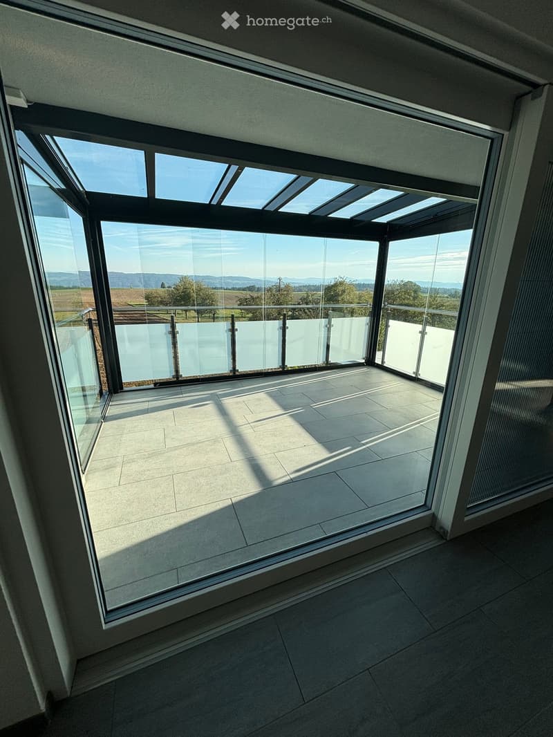 Modern balcony with large glass windows and a tiled floor, offering a scenic view of a rural landscape.