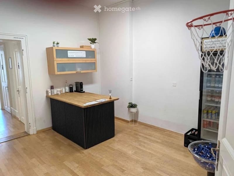 A small break room with a wooden counter, coffee maker, small refrigerator, and a cabinet with glass doors. There are potted plants and a small basket on the floor.