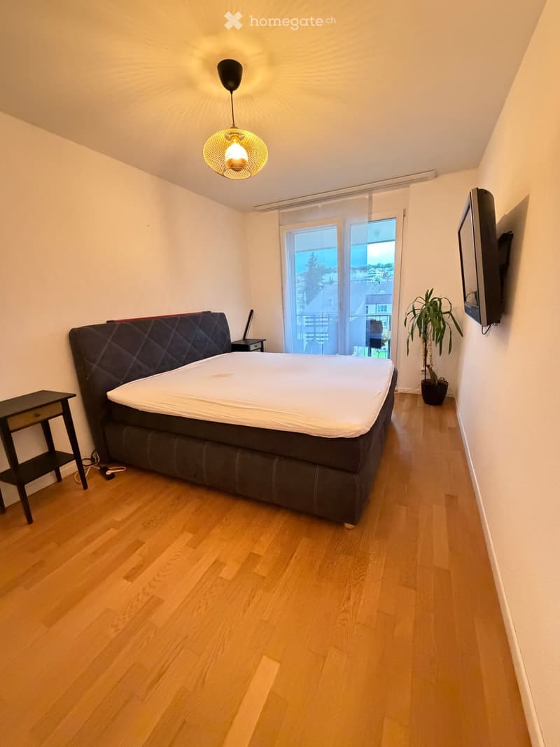 Modern bedroom with a large bed, nightstand, potted plant, and wall-mounted TV. The room features wooden flooring and large windows with a view.