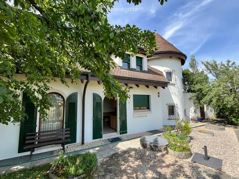 Two-story house with white walls, green shutters, and a red-tiled roof. The house features a round tower with a conical roof and a balcony. The surrounding garden includes a stone bench, potted plants, and a circular stone planter.