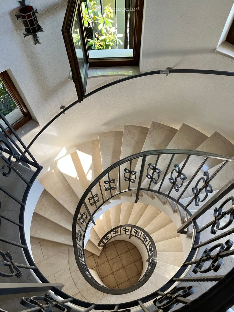 Spiral staircase with beige marble steps and ornate black wrought-iron railing, viewed from above, with a window showing greenery outside.