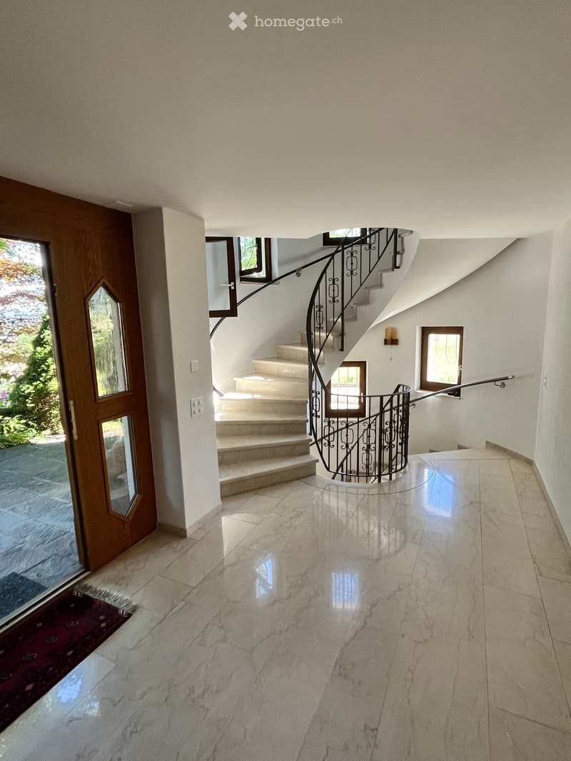 Spacious hallway with polished marble flooring, a curved staircase with a black wrought-iron railing, and large windows allowing natural light to enter. A wooden door leads outside, and a small red rug is placed near the entrance.
