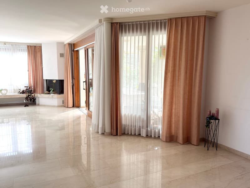 Bright, spacious living room with large windows and sliding glass doors leading to a balcony. Features include a polished marble floor, light-colored walls, and a small side table with candles and a plant. A TV screen is mounted on the wall near the windo