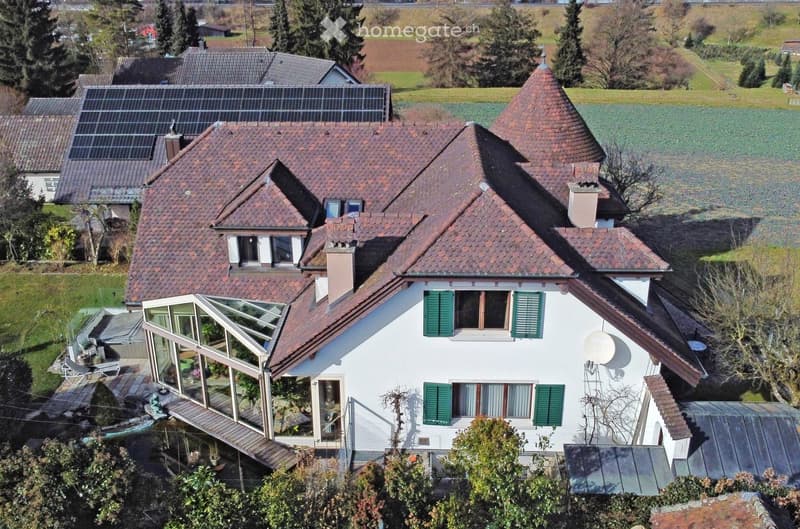 A two-story house with a red-tiled roof, white walls, green shutters, and a large glass sunroom attached to the side. The house features solar panels on the roof and is surrounded by a well-maintained garden and open fields.