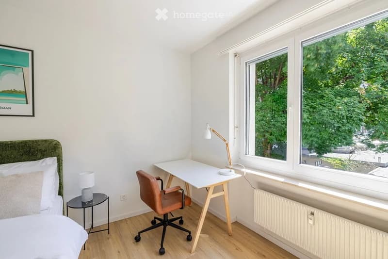 Cozy bedroom with a bed, desk, chair, table lamp, nightstand, and picture frame. Large windows offer a view of greenery outside.