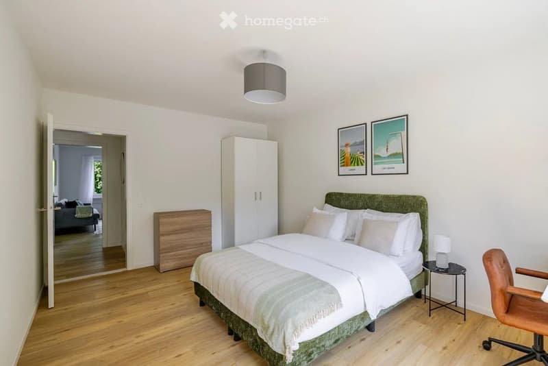 Cozy bedroom with a large bed, nightstand, chair, wardrobe, and a view into an adjacent living space.