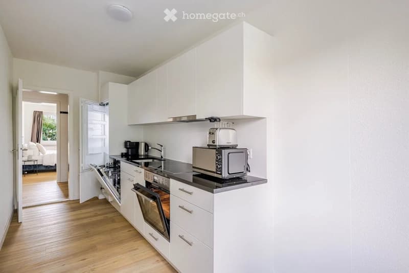 Modern kitchen with white cabinets, stainless steel appliances, and a black countertop. Visible appliances include a refrigerator, dishwasher, oven, microwave, toaster, and coffee maker. The kitchen features a sink with a faucet and is connected to a bedr