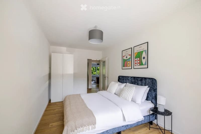 A modern bedroom with a large bed, white bedding, a nightstand with a lamp, a wardrobe, and two abstract picture frames on the wall. The room has wooden flooring and a view to another room through an open doorway.