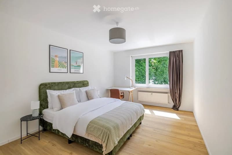 Cozy bedroom with a green upholstered bed, white bedding, nightstand, desk with chair, and large windows with curtains.