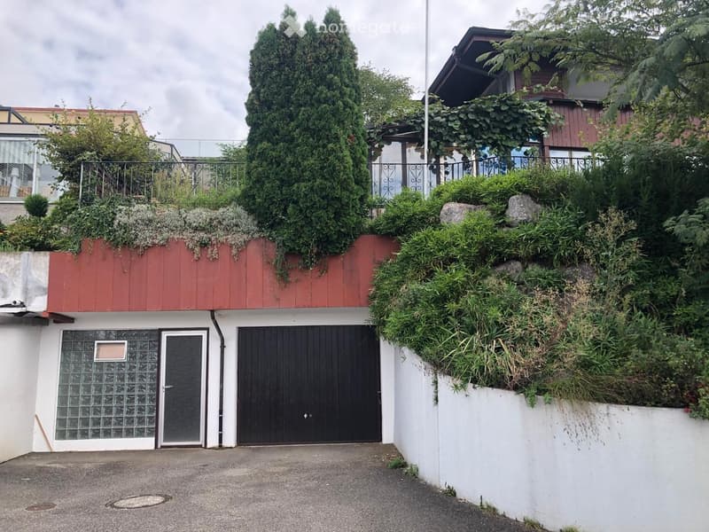 Two-story building with a red roof, white walls, and a black garage door. The building has a landscaped rooftop with greenery and a metal railing.
