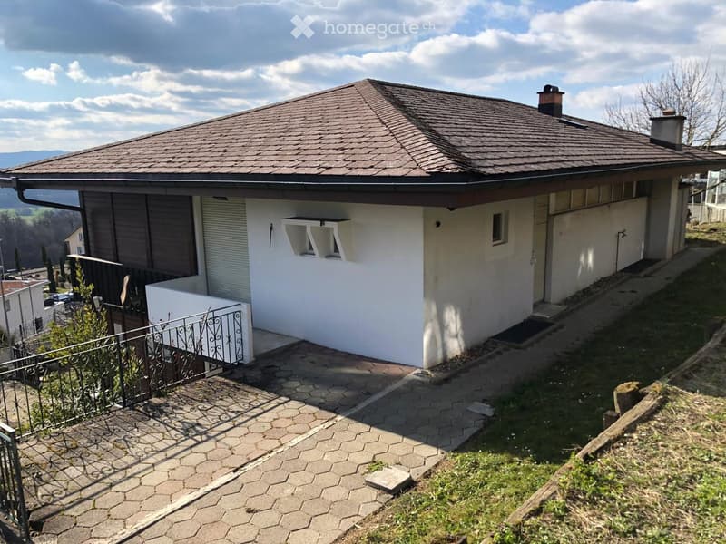 Two-story house with a tiled roof, white exterior, and a small balcony area with a decorative metal railing. The house is situated on a raised ground floor with a paved pathway leading to the entrance.