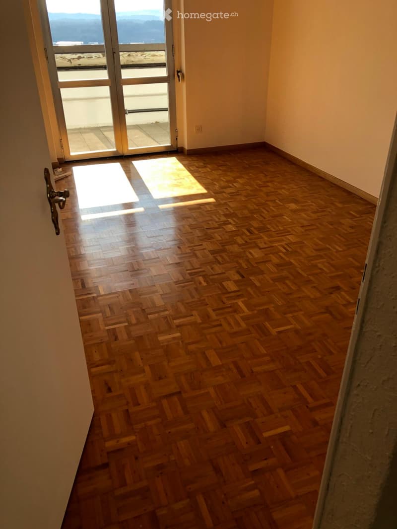 Empty room with wooden herringbone flooring, large glass sliding doors leading to a balcony with a scenic view.