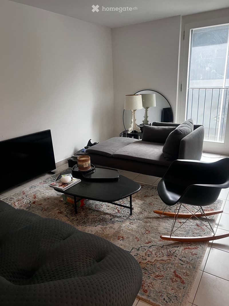 Modern living room with a grey sofa, black coffee table, rocking chair, and a large mirror. The room features a balcony with a view and a television on the left side.