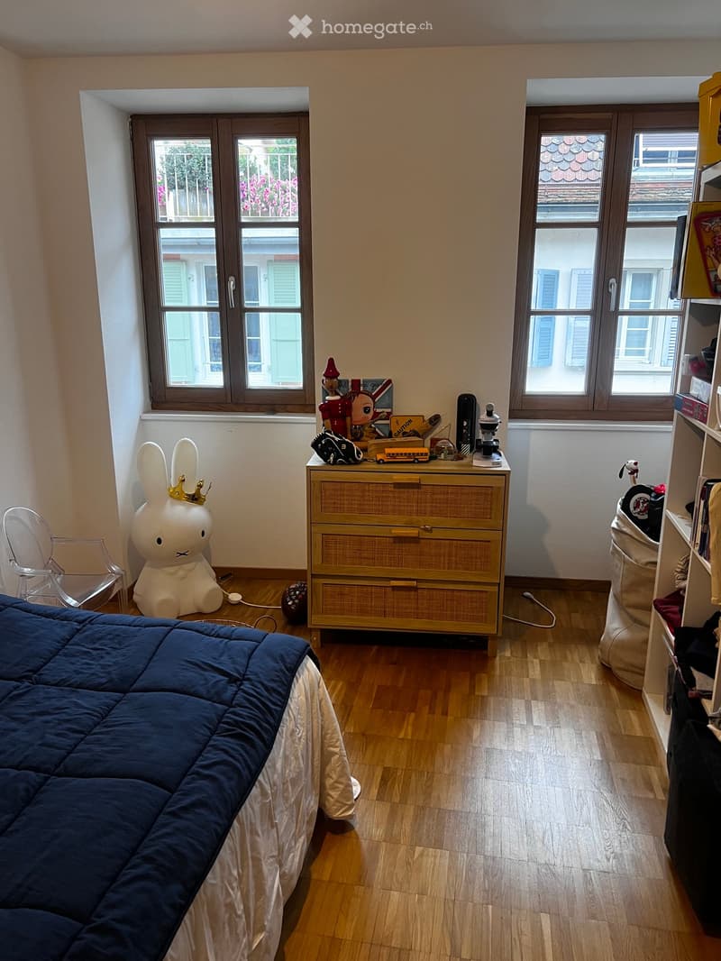 A well-lit bedroom with wooden flooring, a large bed with dark blue bedding, a transparent chair, a large decorative rabbit, a wooden drawer unit with toys and books on top, and a shelf with various items.